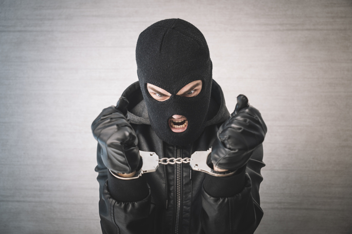 a bandit in black mask and gloves with handcuffs in his hands on a white background. Release from imprisonment. an evil thief is trying to break the chain on the handcuffs.