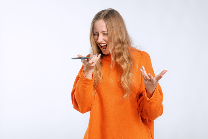 Angry woman arguing on call, screaming at mobile phone speakerphone, recording voice message with furious face, standing in an soft orange sweater over white background