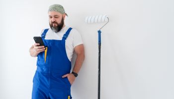 A blue-collar man standing at the wall with a paint roller and using smartphone