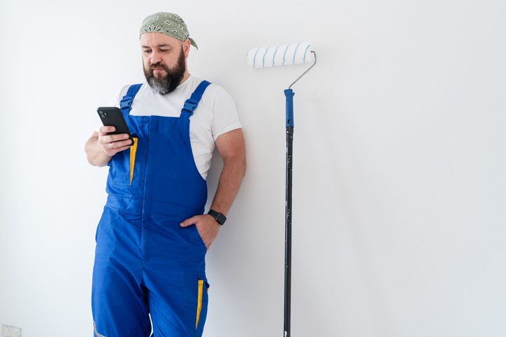 A blue-collar man standing at the wall with a paint roller and using smartphone