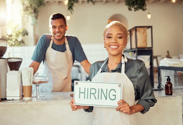 Portrait, leader and hiring sign small business owner happy at coffee shop or cafe with employee. Team, collaboration and recruitment due to startup growth and vacancy to join us for a job